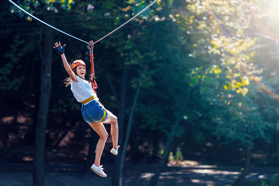 Adventure and Entertainment Insurance - Cheerful Woman Waves as She is Gliding Along a Zipline in an Adventure Park